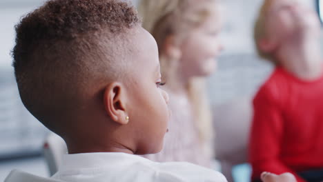 Close-up-of-Black-schoolboy-sitting-on-chair-in-infant-school-talking-to-his-classmates,-back-view