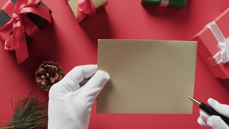 Video-of-santa-claus-holding-brown-envelope-with-copy-space-and-christmas-presents-on-red-background