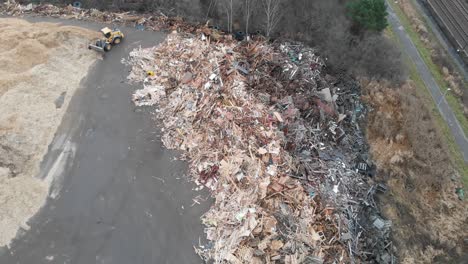 Aerial-rises-in-helix-movement-above-huge-dumpsite-filled-with-tons-of-wooden-and-bulky-waste-while-yellow-tractor-shovels-sawdust