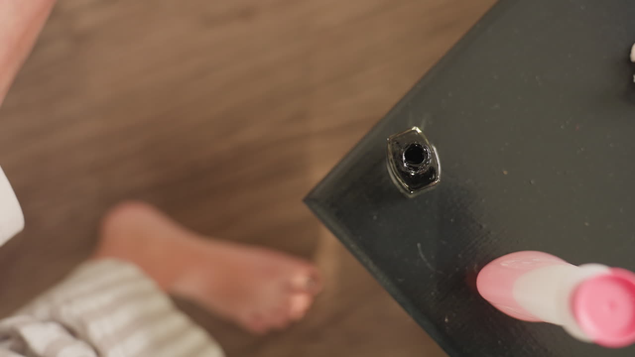 Aerial view shows female hands twisting white cap, dipping brush into black nail polish bottle on dark table, lifting brush out for pedicure session, pink remover container beside