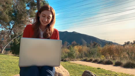 A-happy,-attractive,-young,-caucasian-woman-on-her-laptop-at-the-park,-sitting-on-a-rock---she-smiles-and-lets-out-a-little-laugh