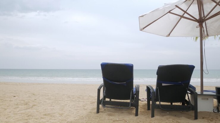 Lussuosi lettini da spiaggia con ombrellone su sabbia bianca con palma da cocco in Thailandia, con clima estivo soleggiato e cielo azzurro limpido