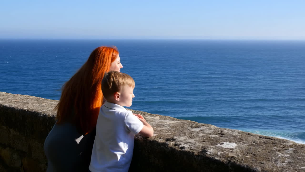 Donna e bambino, oceano blu, muro di pietra, capelli rossi, luce solare, onde calme, orizzonte, intimo legame madre-figlio