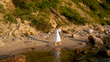 Aerial-View-Of-Women-Wearing-Flowing-White-Dress-Walking-Along-Rocky-Beach-Coastline-During-Golden-Hour