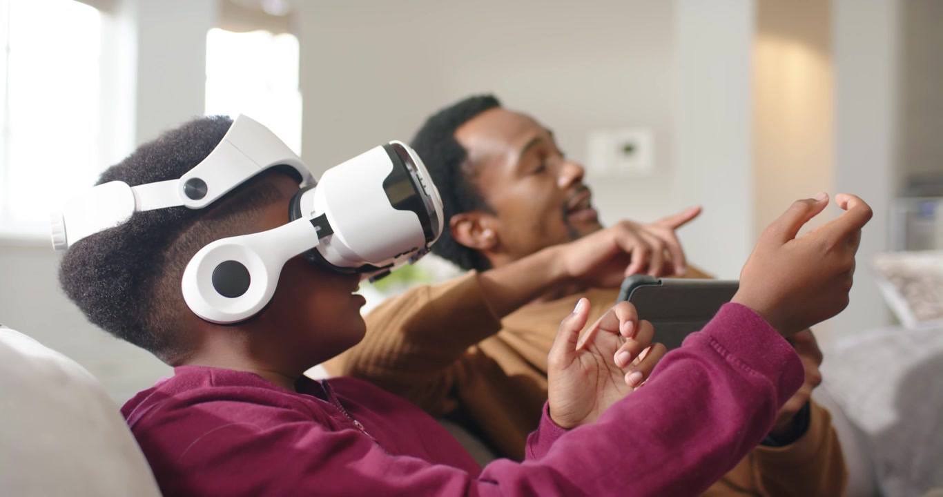 African American dad holding tablet pointing guiding teen with white VR headset gesturing on couch