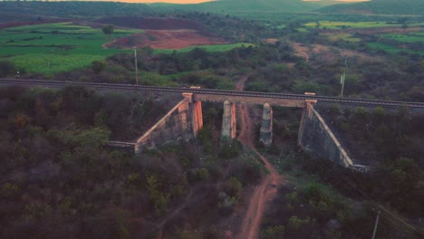 Toma-Aérea-De-Un-Antiguo-Puente-Ferroviario-De-Hormigón-Con-Vías-Férreas-Con-Densas-Colinas-Forestales-En-El-Fondo-Durante-La-Tarde