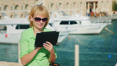 A-Female-Tourist-Enjoys-The-Tablet-Against-The-Backdrop-Of-The-Bay-With-Yachts