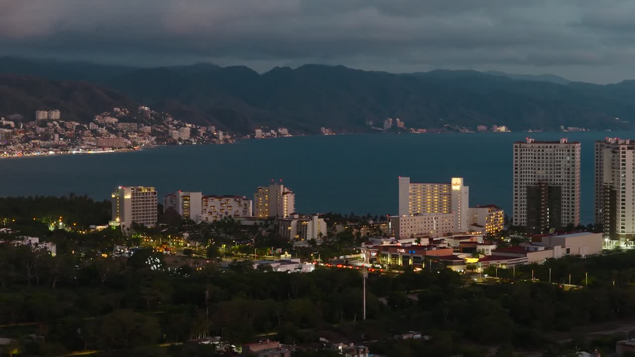 Hotels and high-rises illuminated along the Puerto Vallarta coastline with the Pacific Ocean and Sierra Madre mountains at dusk