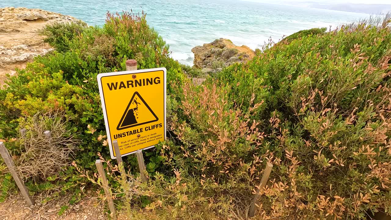 A warning sign highlights unstable cliffs near lush greenery and ocean views at Aireys Inlet, Victoria, Australia
