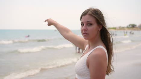 Chica-En-Bikini-Blanco-Mira-A-La-Cámara-Mientras-Señala-El-Mar-Y-Asiente-Con-La-Cabeza-En-La-Playa-En-Verano