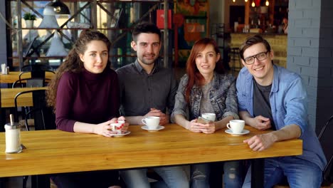 portrait of four young people friends in casual clothes sitting at table in spacious cafe with tea cups and looking at camera. eating out and friendship concept.