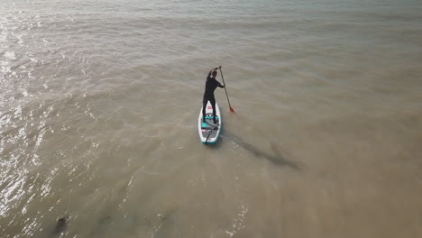 Young-man-hops-up-onto-paddle-board-in-the-sea-with-boat,-glistening-water-and-blue-sky-in-the-background