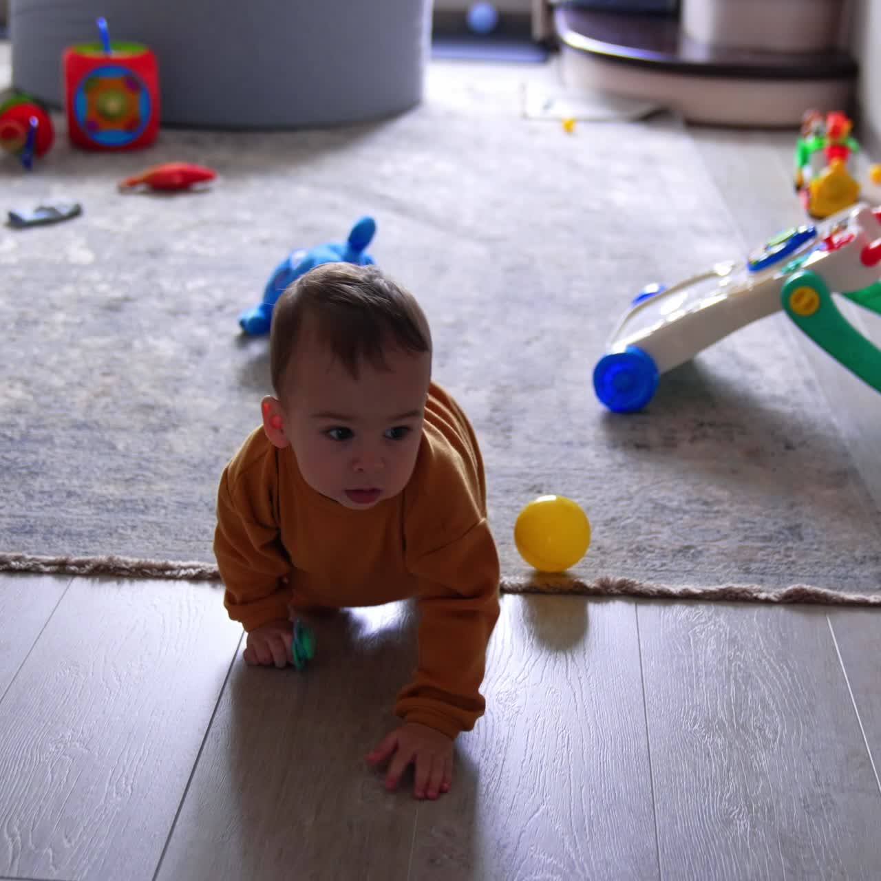 Pretty toddler in orange sport suit crawls by the floor indoors. Bright toys on the carpet in the room at backdrop