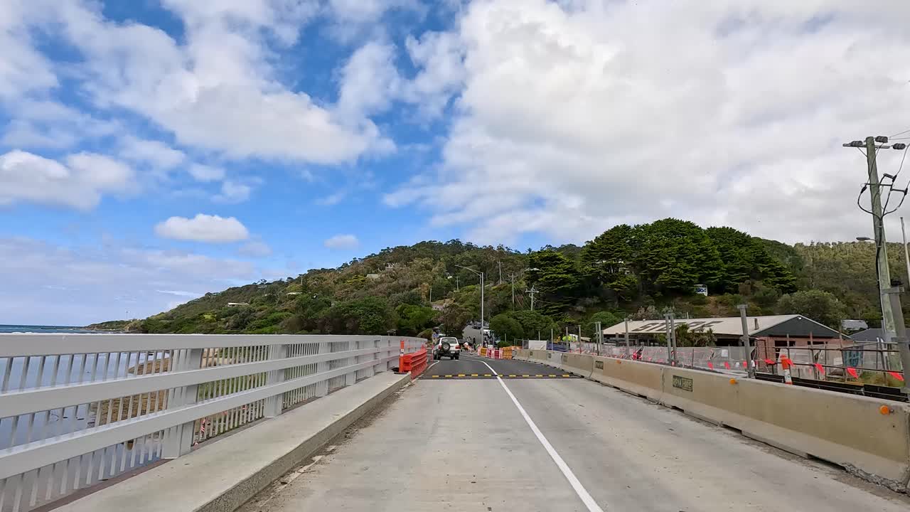 A vehicle travels across a bridge on Great Ocean Road, surrounded by scenic hills and blue skies, under bright daylight