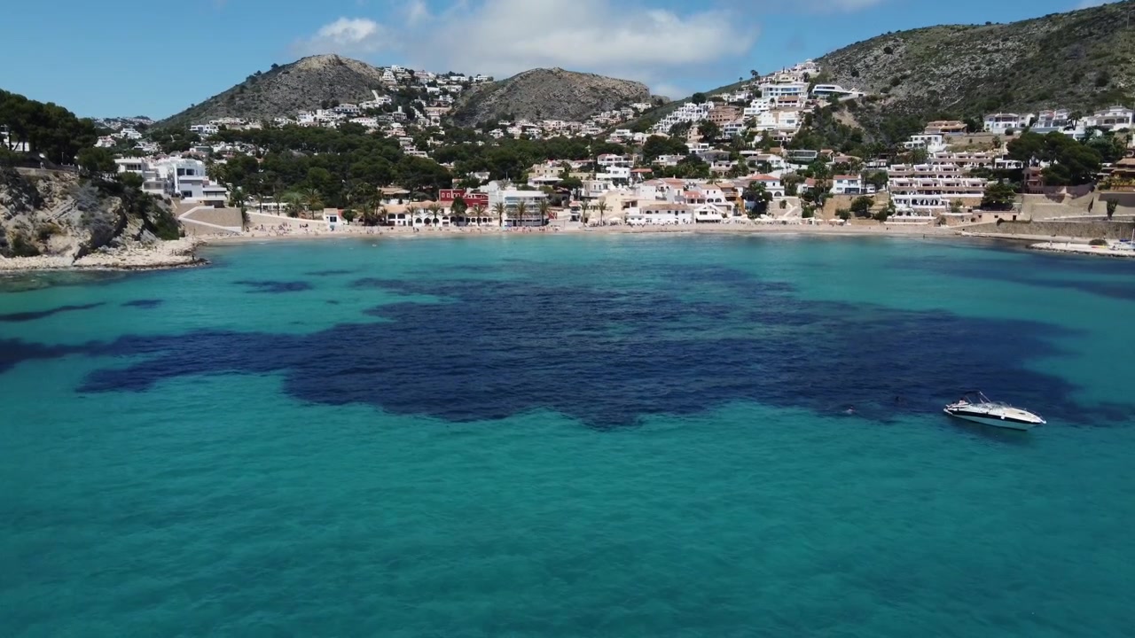 Drone descendo baixo sobre o mar calmo em direção a um veleiro solitário ancorado, com a costa cénica e a praia de El Portet, Moraira, Espanha, ao fundo
