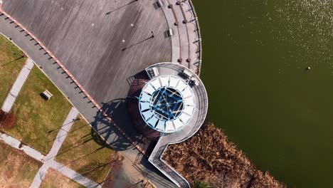 a top down shot over a circular building next to a lake on a sunny day in a park