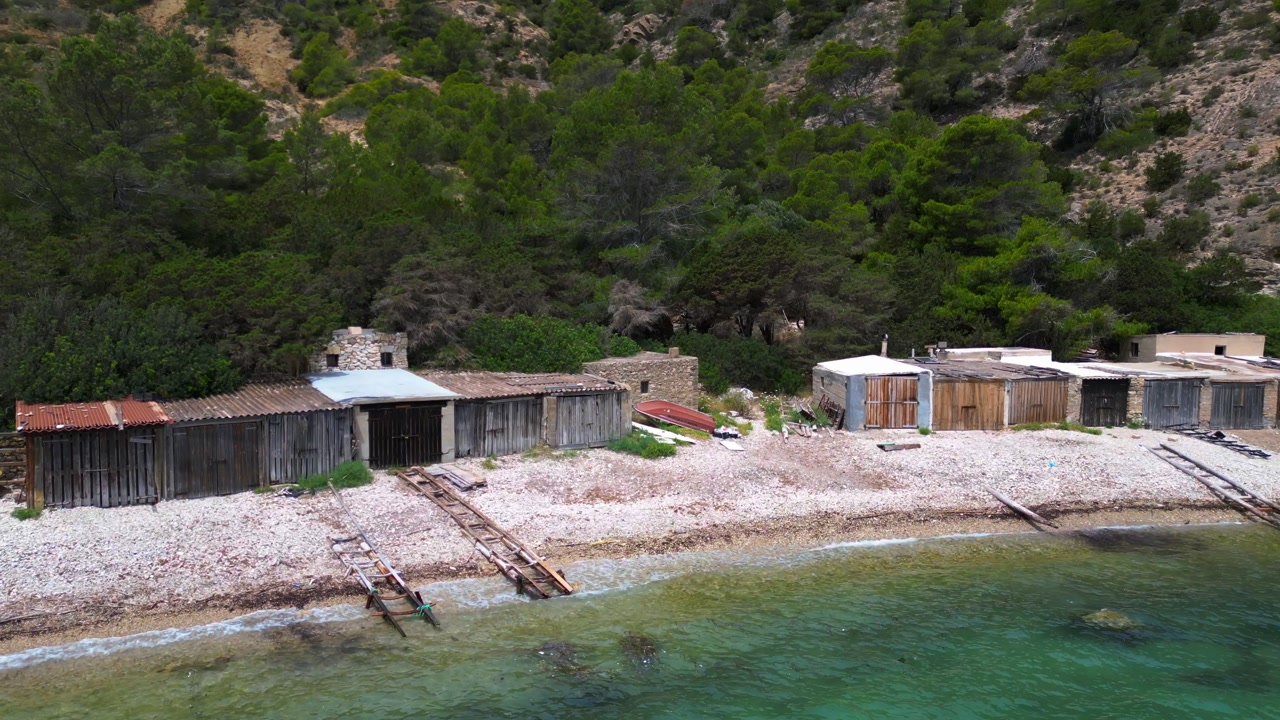 Cala Llentrisca, a secluded beach in Ibiza with Historic boat garage, highlighting the pristine turquoise waters and surrounding verdant landscape. Nice aerial view flight fly push forward drone