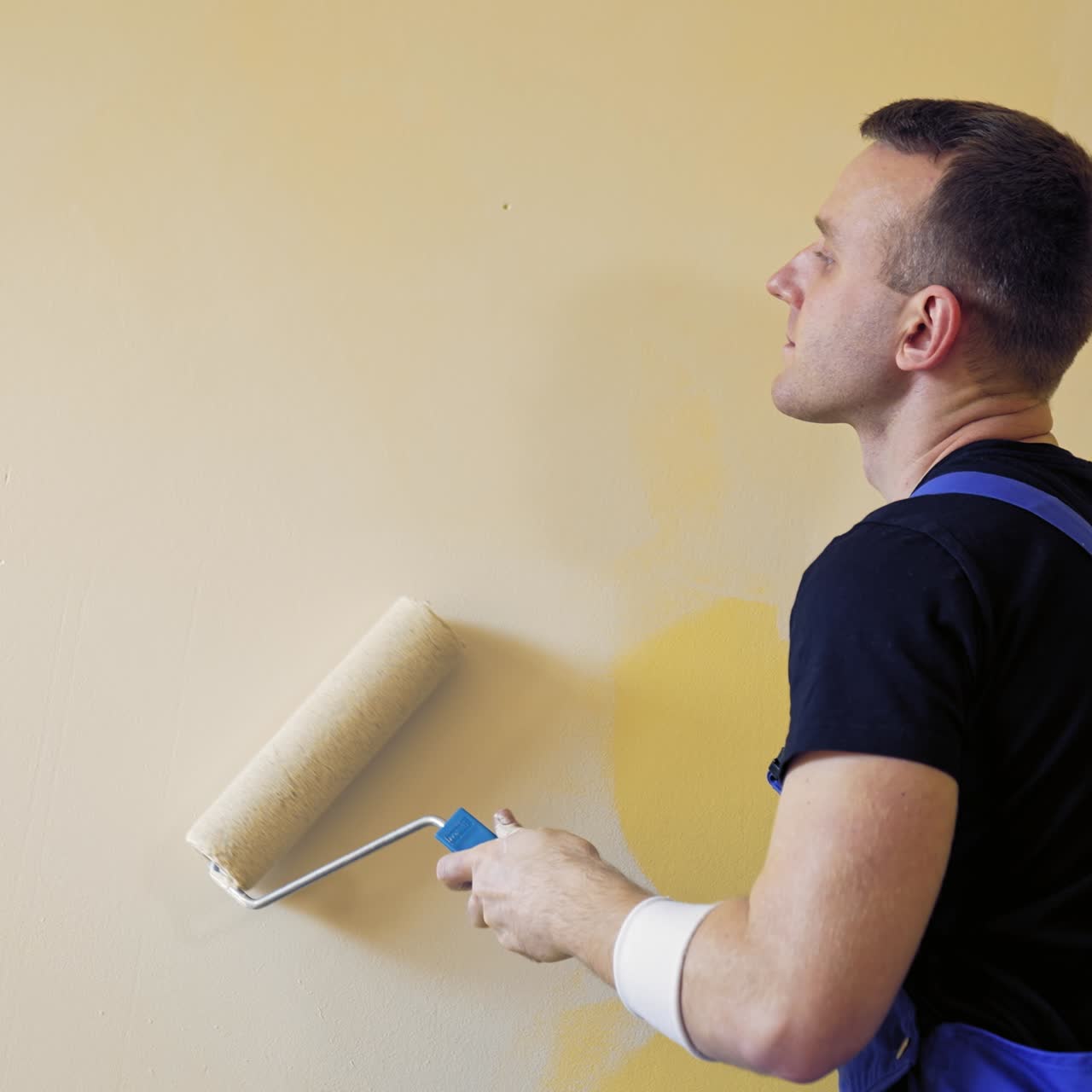 Operaio con un rullo per pittura. Giovane uomo in tuta da lavoro che dipinge una parete di colore chiaro. Servizi di pittura professionali.