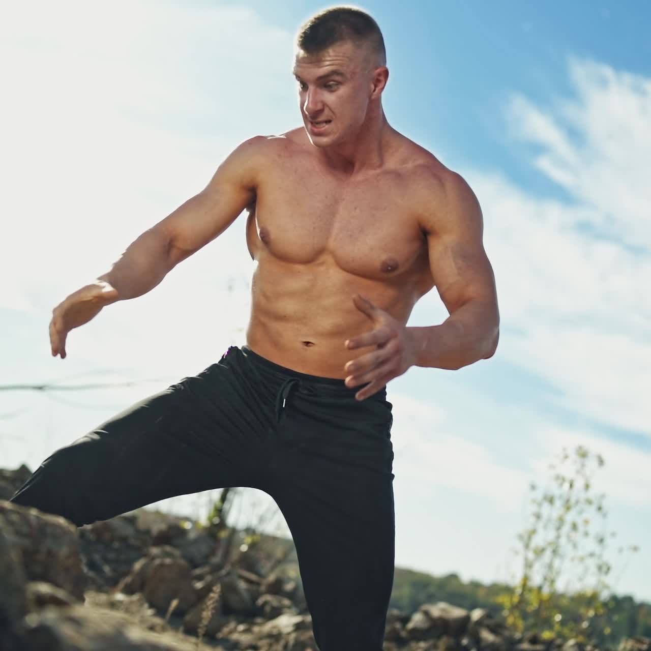 Muscular man with trained body exercises with stones. Strong bodybuilder doing workout by throwing heavy rocks on nature background.