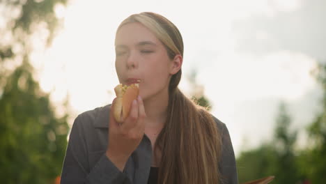 lady wearing grey top enjoys eating burger with a look of satisfaction, eyes closed, in a sunlit outdoor setting, background blurred with sunrays illuminating scene