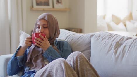 video of thoughtful biracial woman in hijab drinking coffee relaxing on sofa in living room at home