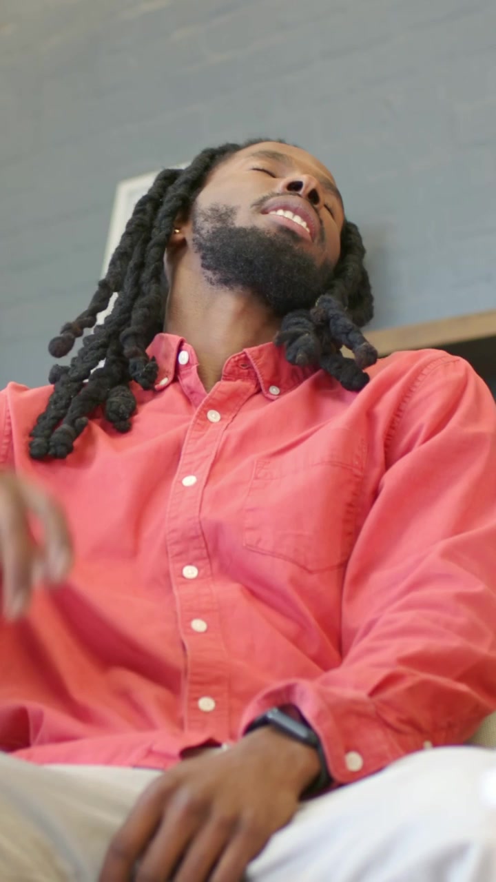 Vertical video: Sighing, man with braids in coral shirt pressing brow in woven armchair, resting