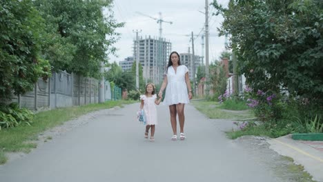 a pregnant woman and her young daughter, dressed in light summer outfits, walk hand-in-hand along a street in a residential neighborhood. the daughter holds a toy, enjoying the peaceful moment outdoor