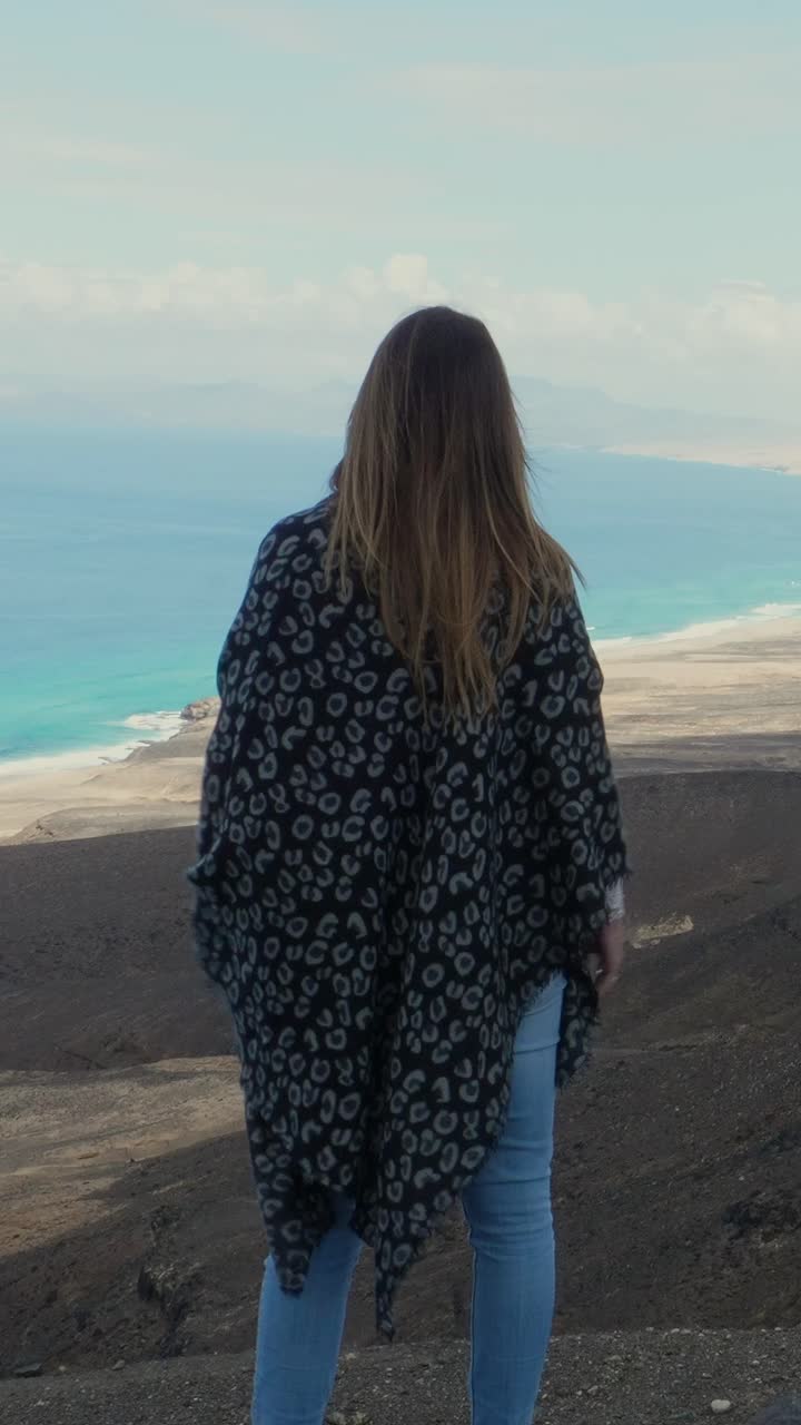 Woman from behind standing on top of a mountain watching the ocean with a scarf in the wind