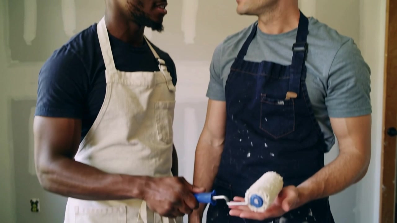 Presenting man in splattered apron showing paint roller to partner in reno room, preparing walls