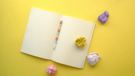 libro de notas abierto con bolígrafos y papeles arrugados en fondo amarillo