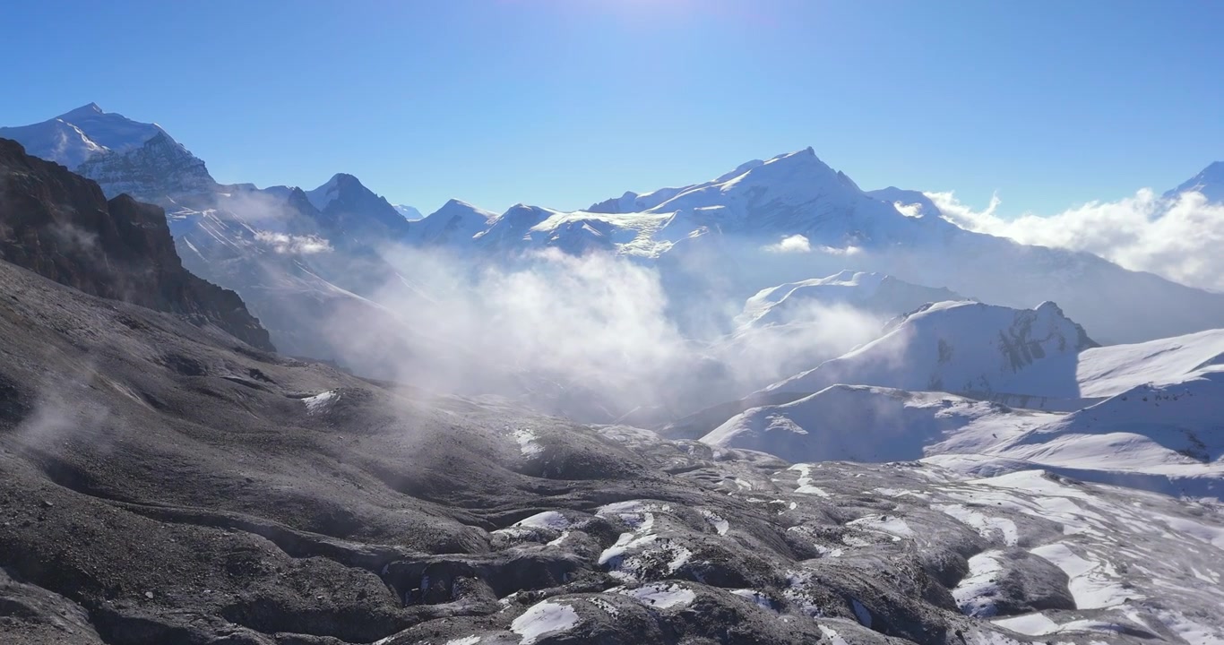 Annapurna Circuit, Manang, Nepal, droneoptagelse af skyer der driver over barskt alpint terræn med sneklædte Himalaya-bjergtoppe i skarpt morgenlys på ruten til Thorong La Pass