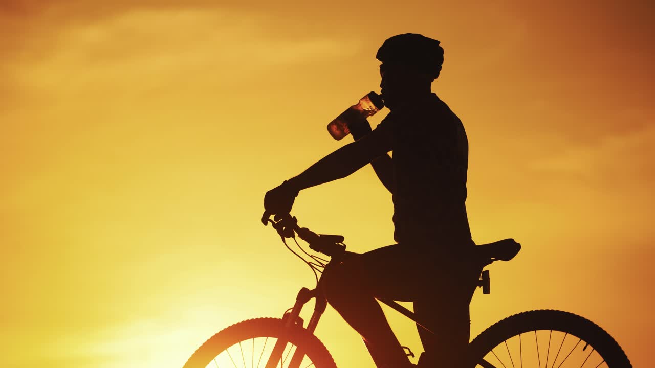 Silhouette of professional bicyclist taking a bottle and drinking water at golden sunset. Male cyclist in helmet drinks water on the natural background of setting the sun in the evening.