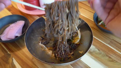 Manos-Mezclando-Fideos-De-Trigo-Sarraceno-En-Un-Tazón-Con-Palillos-De-Acero,-Cocina-Coreana