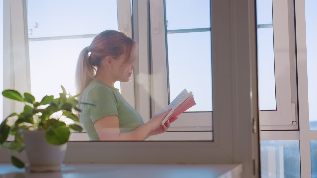Side view young student reading novel near window as she flips to new page, sunlight brightens scene with indoor plant in foreground, symbolizing study, knowledge and relaxation