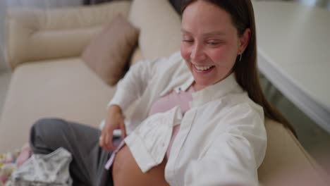 Esta-Mujer-Morena-Embarazada-En-Primera-Persona-Filma-Un-Vlog-Y-Se-Toma-Una-Selfie-Con-Su-Teléfono-Mostrando-Ropa-Para-Su-Pequeño-Hijo-Que-Está-Esperando-En-Casa.