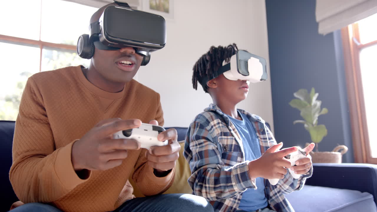 Padre e figlio afroamericani che giocano a un videogioco usando visori VR a casa, al rallentatore.