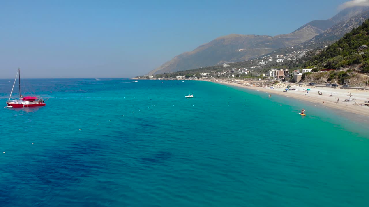 Long sandy beach with umbrellas on shore washed by azure water, anchored boats in Ionian sea