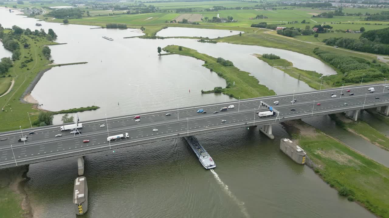 Drone footage of a highway bridge with a barge navigating underneath on a wide Dutch river.