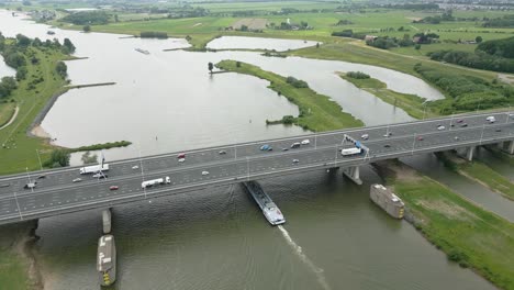 drone footage of a highway bridge with a barge navigating underneath on a wide dutch river.