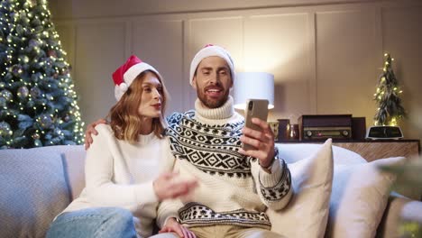 Alegre-Pareja-Familiar-Hablando-En-Video-Chat-En-Línea-En-El-Teléfono-Inteligente-Y-Saludando-Con-La-Mano-Mientras-Se-Sienta-En-Casa-Con-El-Brillante-árbol-De-Navidad-Marido-Y-Mujer-Saludando-A-Amigos-En-Videollamada-Concepto-De-Año-Nuevo