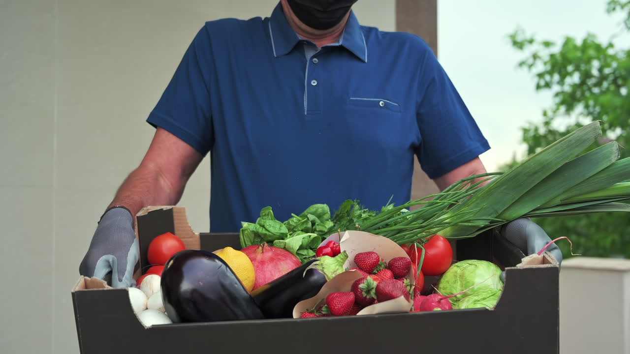 Close up of a man wearing gloves holding a box full of fresh fruits and vegetables