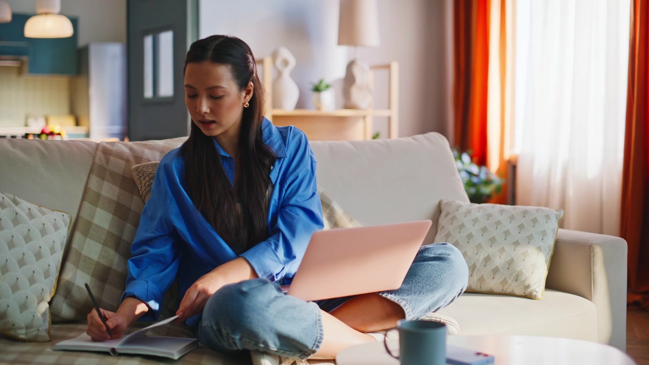 Donna asiatica che lavora al laptop sul divano di casa. Freelance che digita appunti in soggiorno.