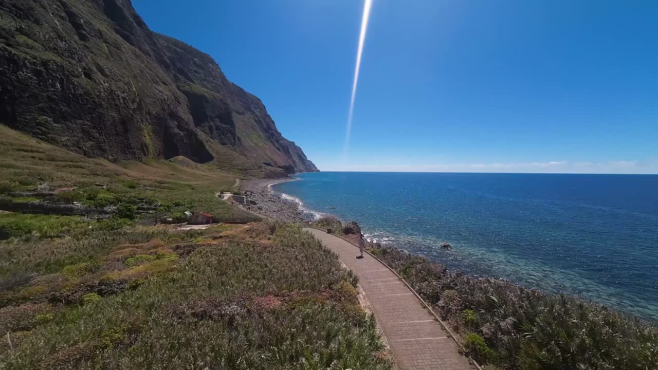 People walk along a brick paver pathway near the sea with steep cliffs rising up from the shoreline. Aerial view from drone of Madeira coast.