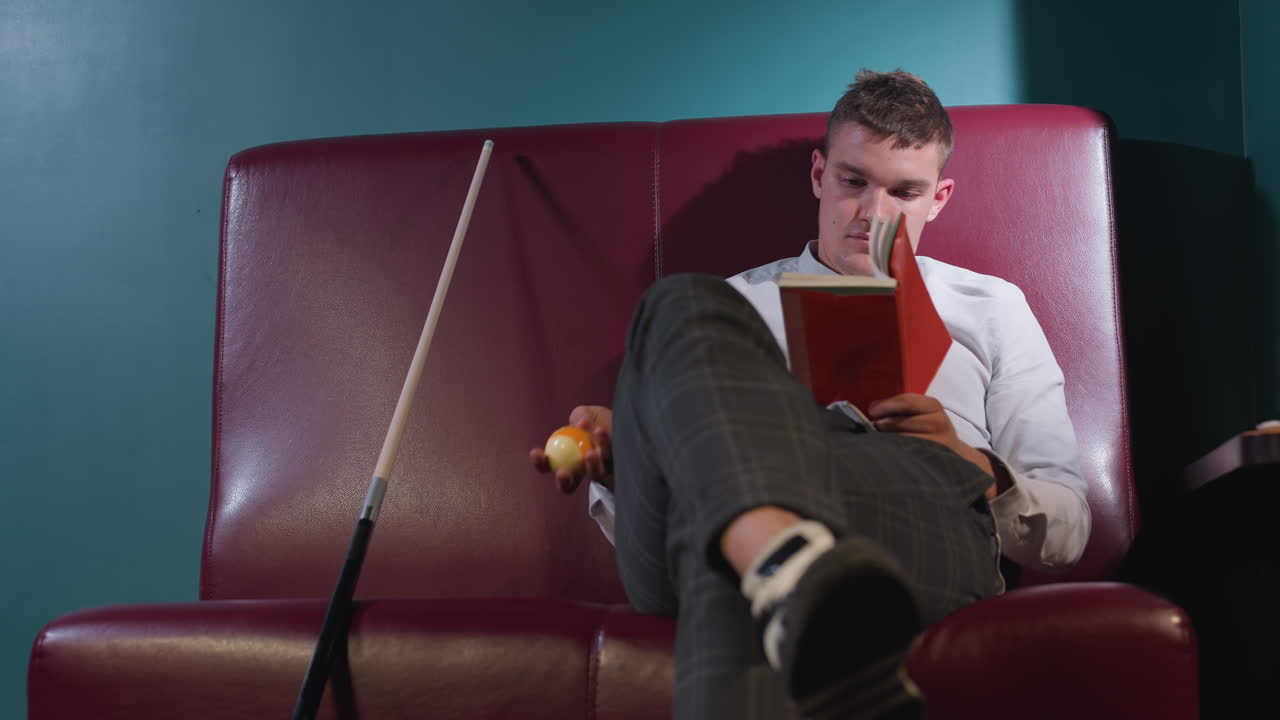 색 셔츠와 칠색 바지를 입은 남자가 빌리어드 방의 빨간 소파에 앉아 다리를 고 오렌지색 빌리어드를 손에 들고 책을 읽고 있다.