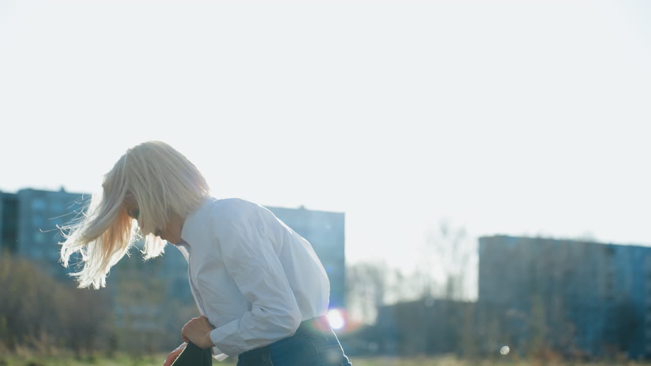 Smiling blonde woman in white shirt outdoors under bright sunlight, hair blowing in breeze, casual and relaxed expression, natural background with soft focus trees and buildings creating lighthearted