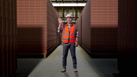 full body of asian male engineer with safety helmet smiling to camera and waving hand saying bye while standing with red brick packed in stacks are stored
