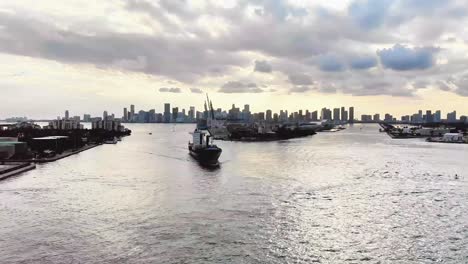 Drone-approaches-Port-of-Miami-with-city-skyline,-passes-closely-over-a-huge-cargo-ship