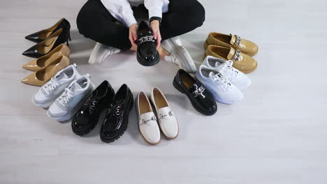 new pairs of modern footwear on the floor in front of sitting lady. woman runs her hands over the diverse shoes choosing one black lacquered loafer. view from above.