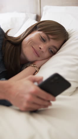 vertical video: caucasian woman lying on bed, holding smartphone at home