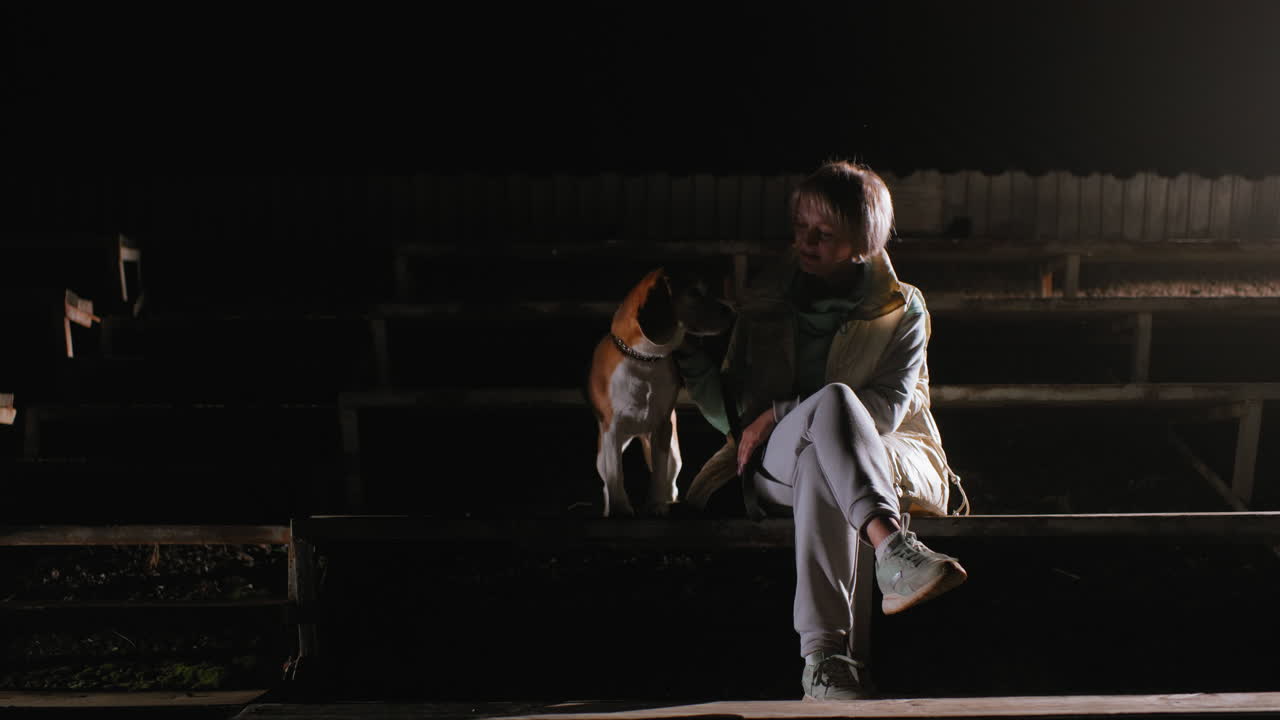Trainer resting beside puppy on podium under dark cloudy night with soft spotlight illuminating scene creating peaceful intimate moment showing trust companionship love in quiet setting